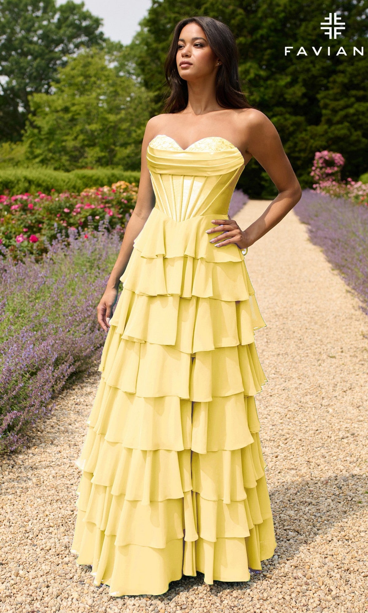 A woman stands on a garden gravel path wearing the Strapless Long Ruffled Prom Gown: Faviana 11436, a yellow tiered dress with a sweetheart neckline. The